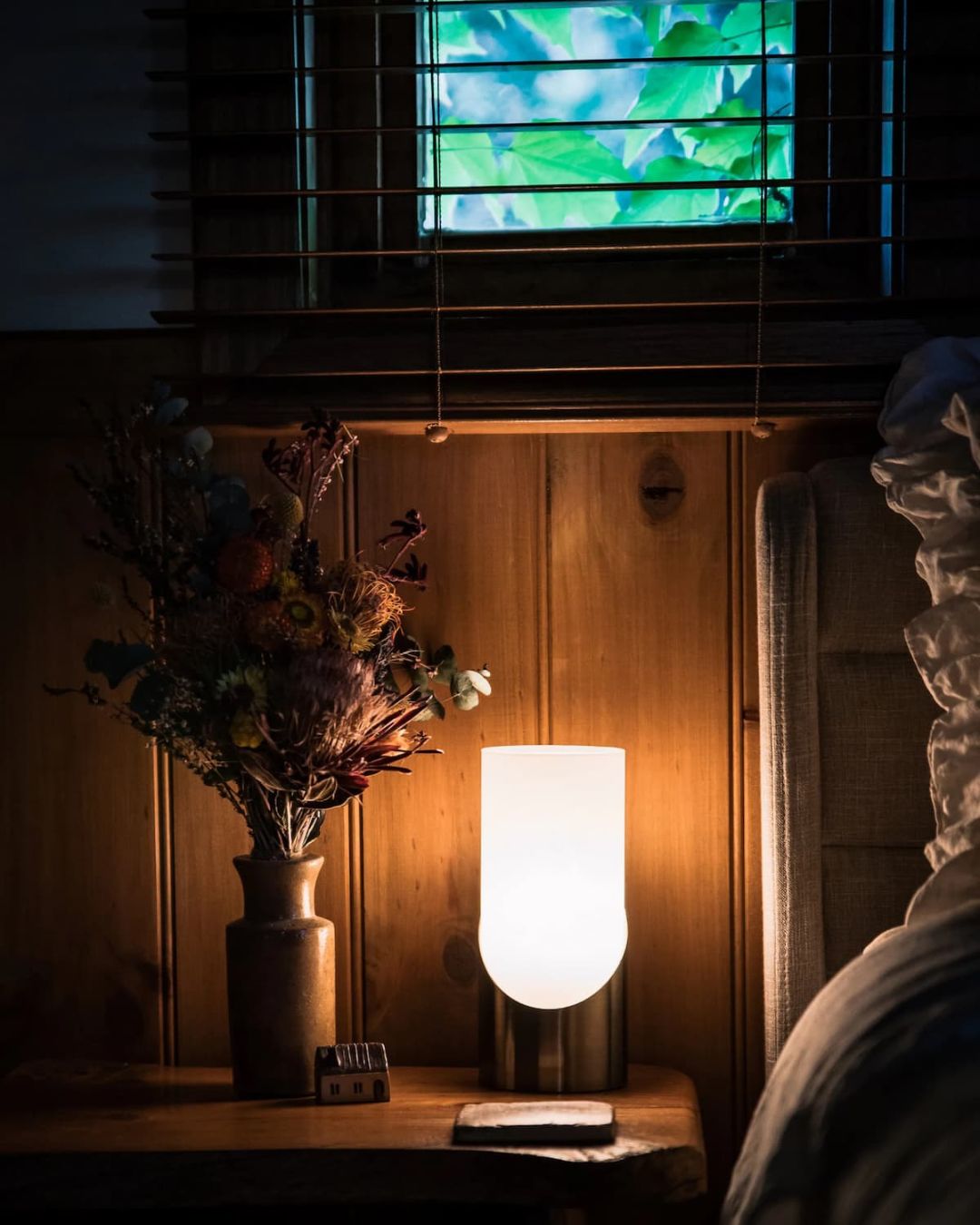 A lit lamp and a vase of dried flowers on a timber sideboard, in warm evening light
