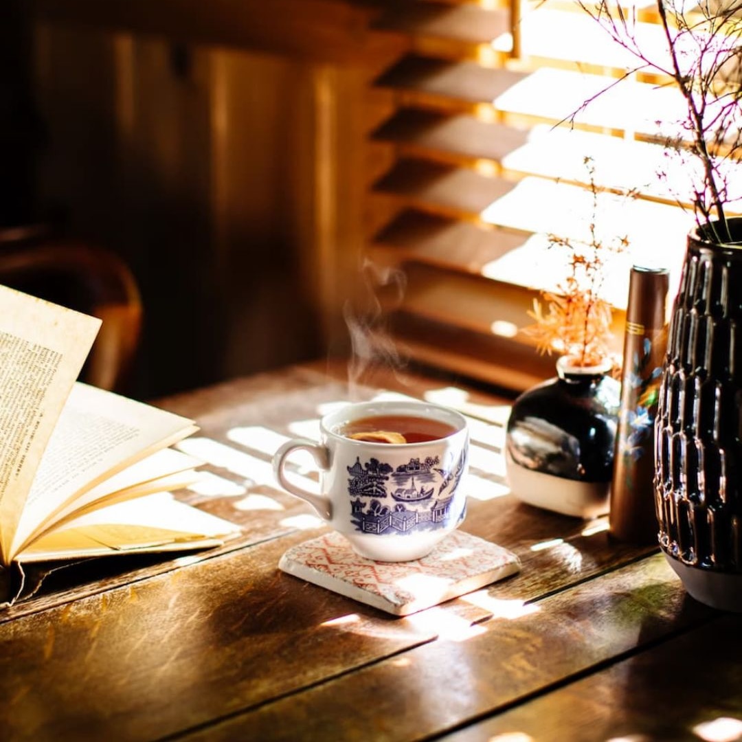 A steaming teacup and an open book on a timber table, with sunlight coming through window blinds and a vase of blossom behind