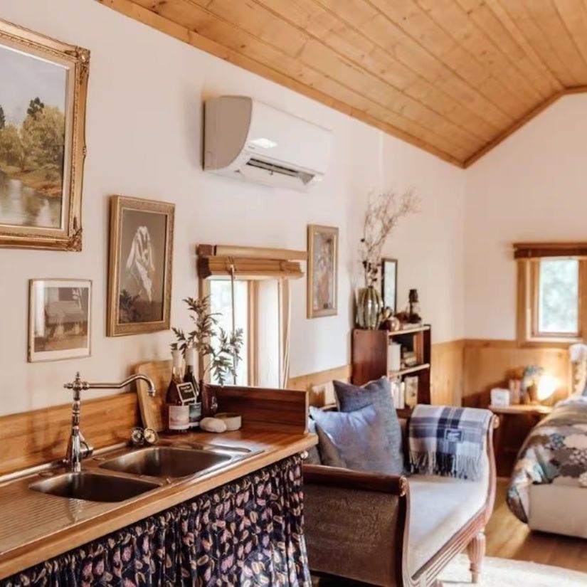 The cottage living space with a timber pitched ceiling, framed pictures on the walls, a leather armchair with a tartan blanket, and a kitchen sink in the foreground