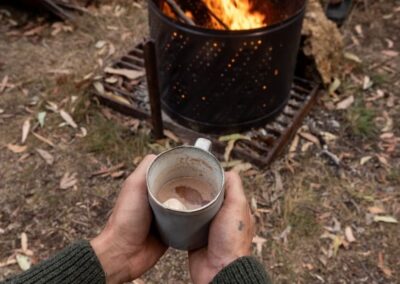 Two hands holding a mug of hot chocolate in front of a lit fire pit