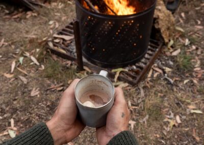Two hands holding a mug of hot chocolate in front of a lit fire pit